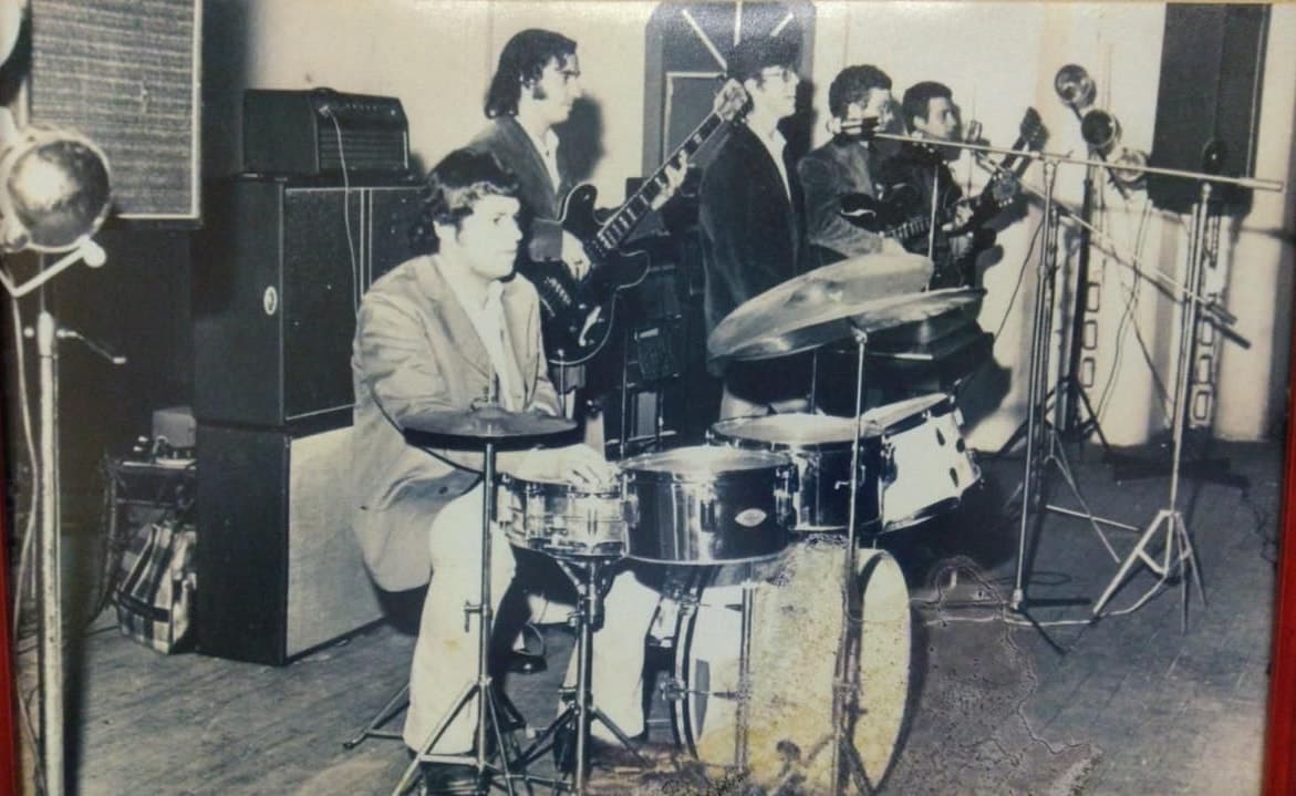 Fotografia em preto e branco de um baile: o baterista de terno à frente, guitarrista e metais atrás dele, caixas de som empilhadas ao lado