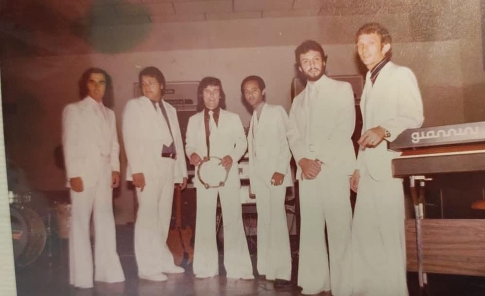 Seis integrantes de terno branco e calça boca de sino em fila no palco, ao lado de um teclado