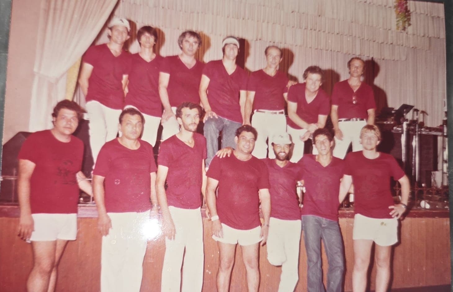 Integrantes de camiseta vermelha posam em duas fileiras à frente do palco, com uma cortina clara ao fundo
