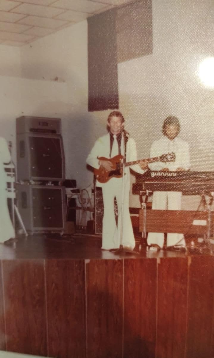 Guitarrista de branco com guitarra vermelha ao lado do tecladista, sobre um palco de madeira com caixas de som