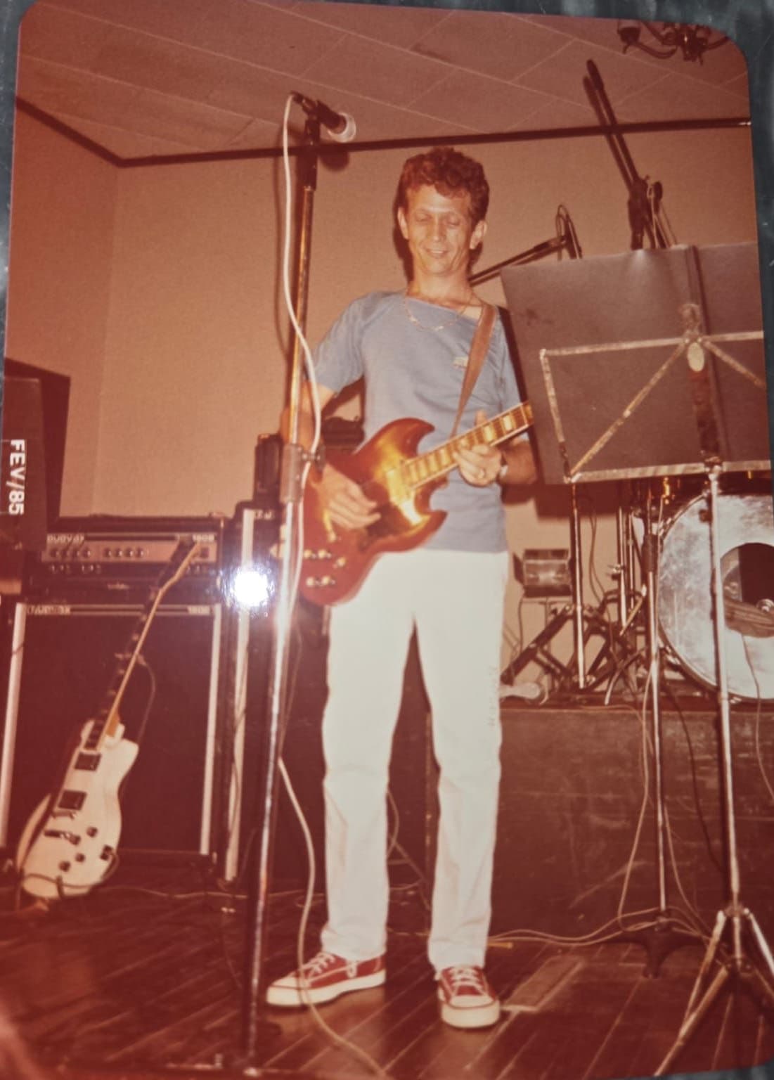 Guitarrista de camiseta azul e calça branca com uma guitarra vermelha, diante de uma estante de partitura; a cópia traz o carimbo FEV/85