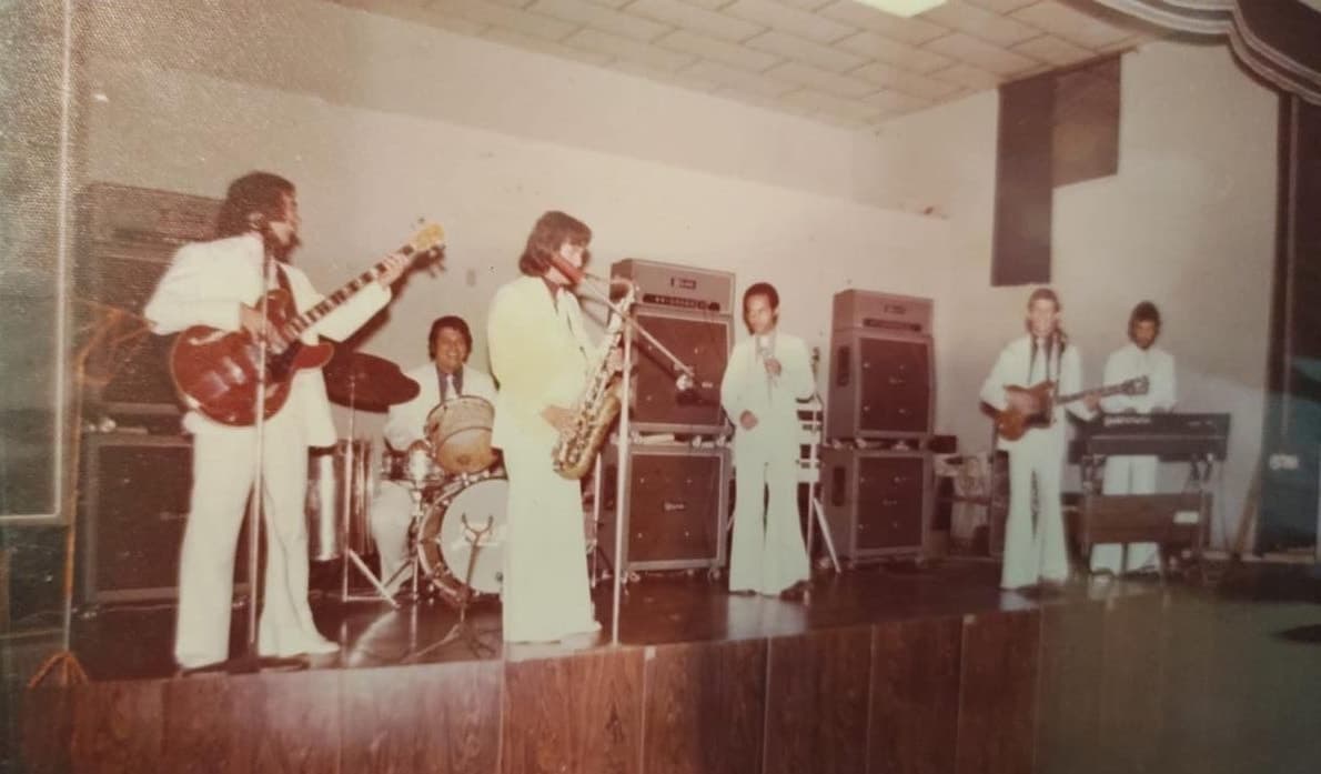 A banda tocando de branco num palco de salão, com guitarra, bateria, saxofone e teclado