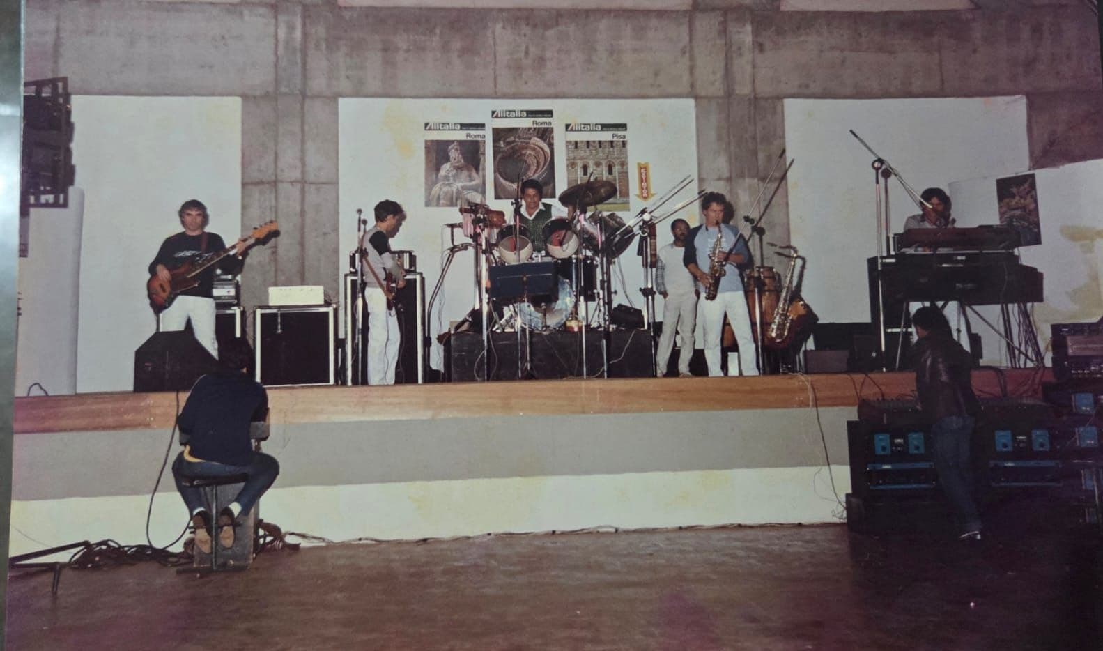 A banda num palco baixo diante de uma parede de concreto com cartazes, vista de trás do técnico de som sentado