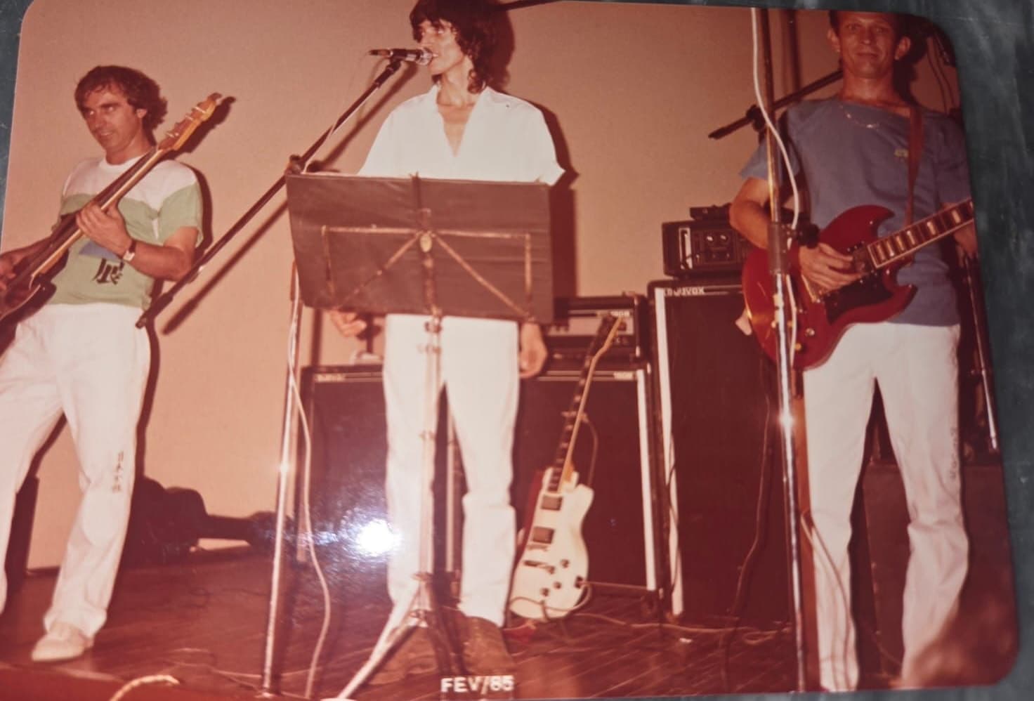 Três músicos lado a lado no palco: dois com instrumentos de corda e, ao centro, o vocalista ao microfone; a cópia traz o carimbo FEV/85