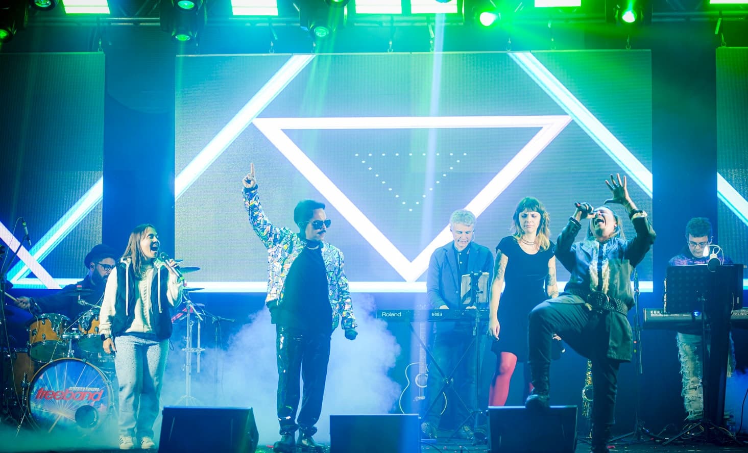 Banda no palco diante do painel de LED com triângulos de luz, vocalistas à frente e teclados e bateria ao fundo