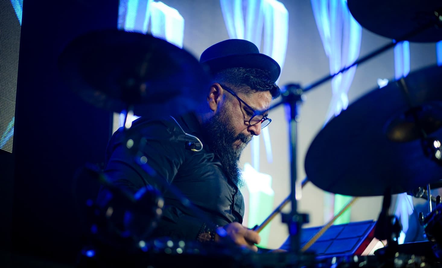 Baterista de chapéu e óculos tocando entre os pratos, com o painel de LED ao fundo