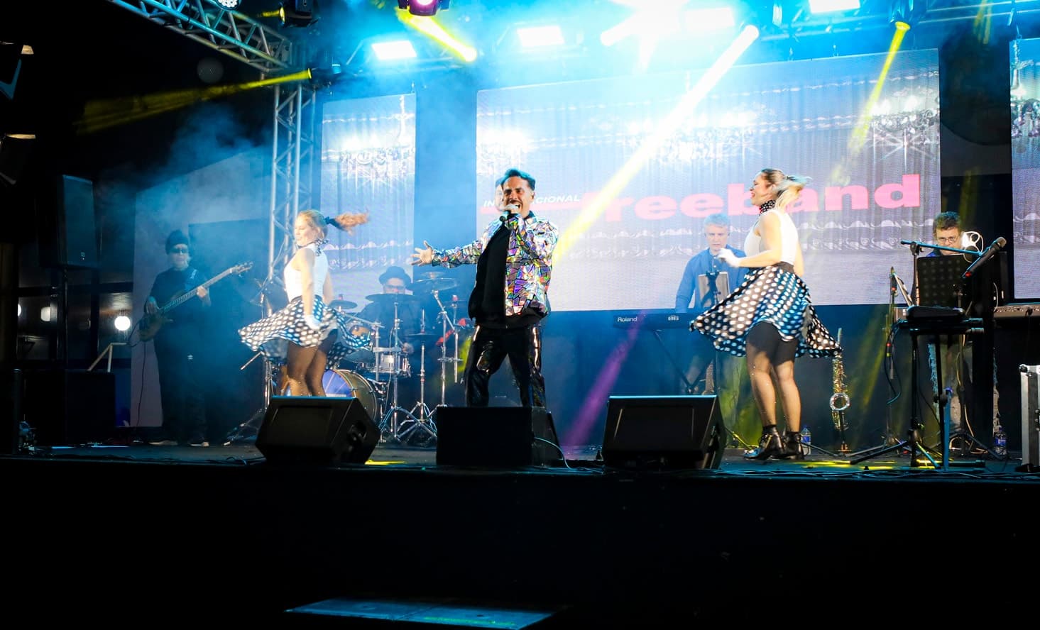Vocalista de jaqueta espelhada entre duas bailarinas de saia de poá rodando, com a banda e o painel de LED ao fundo