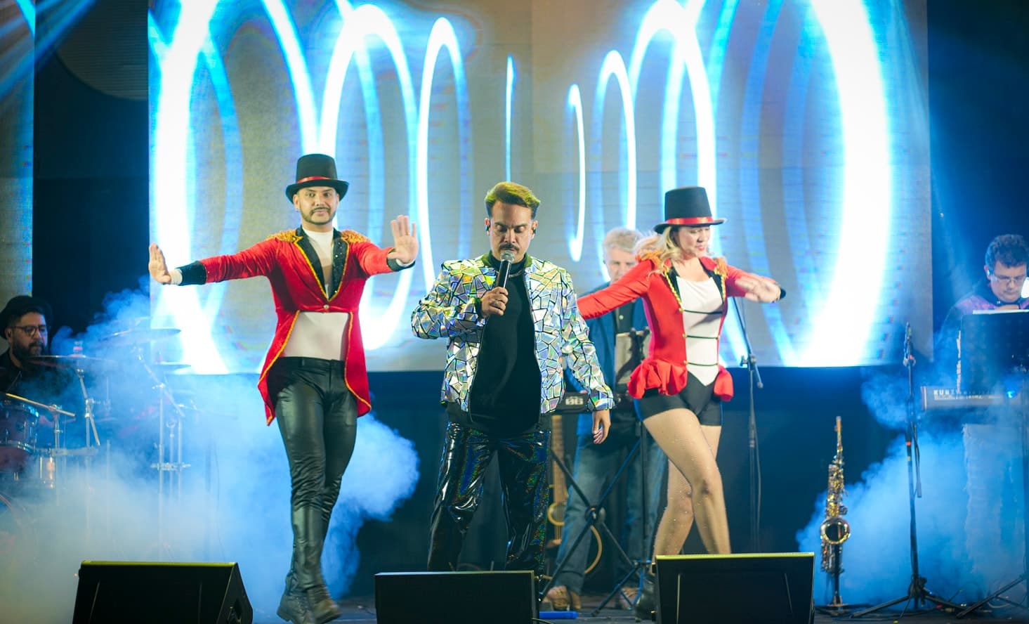 Vocalista de jaqueta espelhada entre bailarino e bailarina de cartola e casaca vermelha, com fumaça no palco