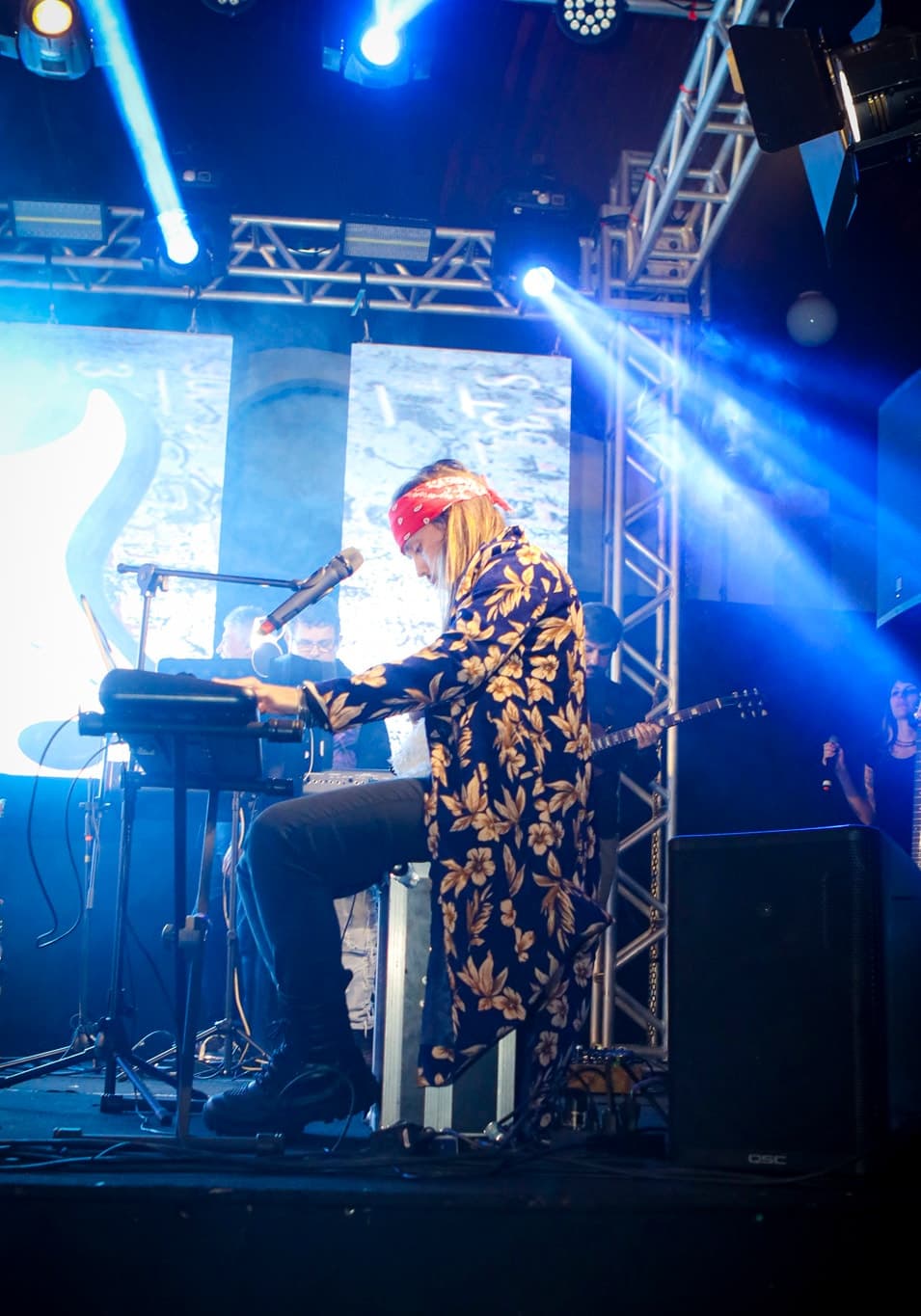 Vocalista de bandana vermelha tocando teclado sob feixes de luz azul e treliças