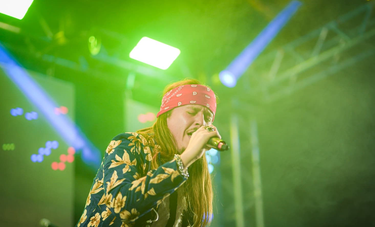 Vocalista de bandana vermelha e camisa floral cantando ao microfone sob luz verde