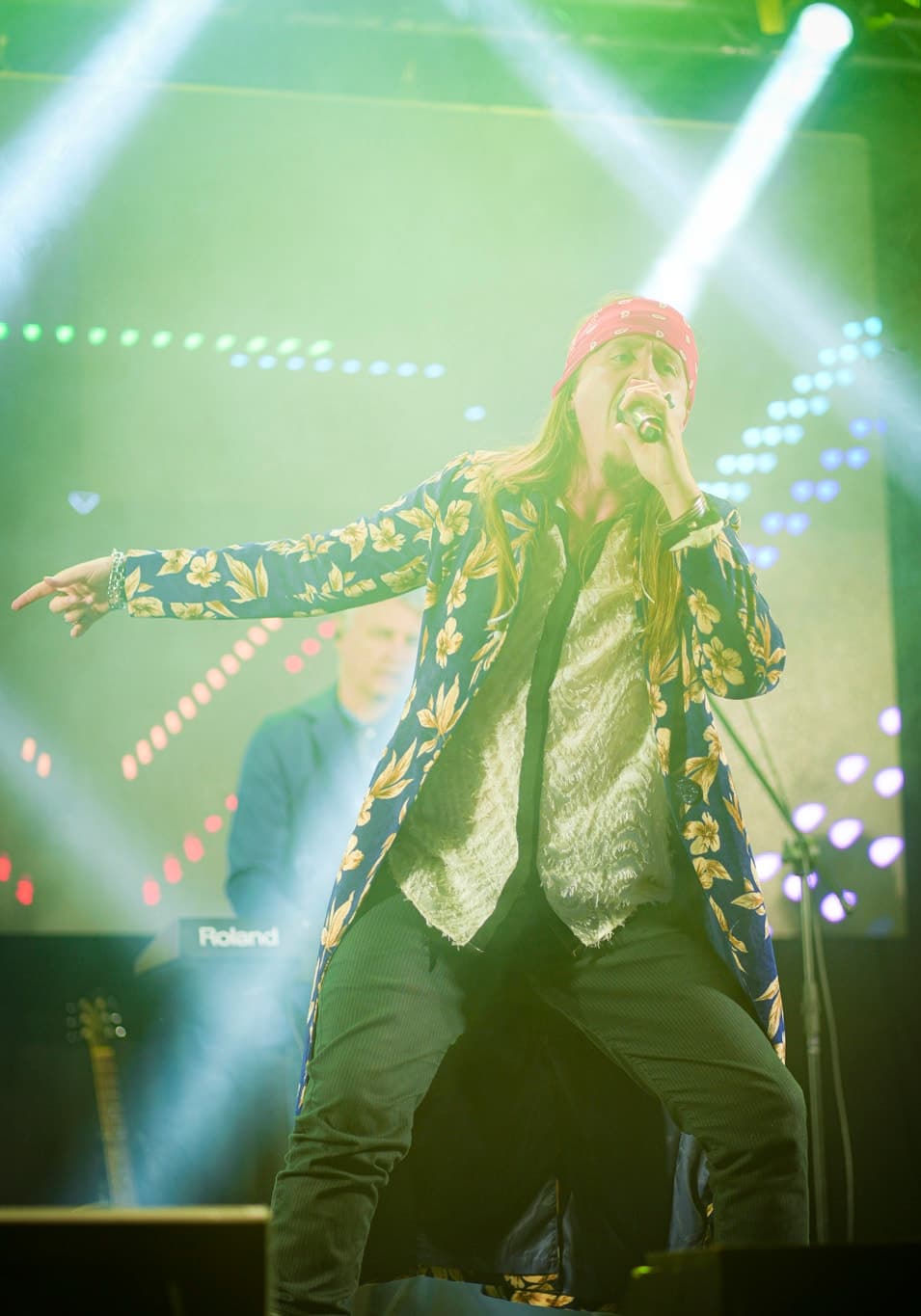 Vocalista de bandana vermelha e casaco floral cantando de braço aberto sob feixes de luz