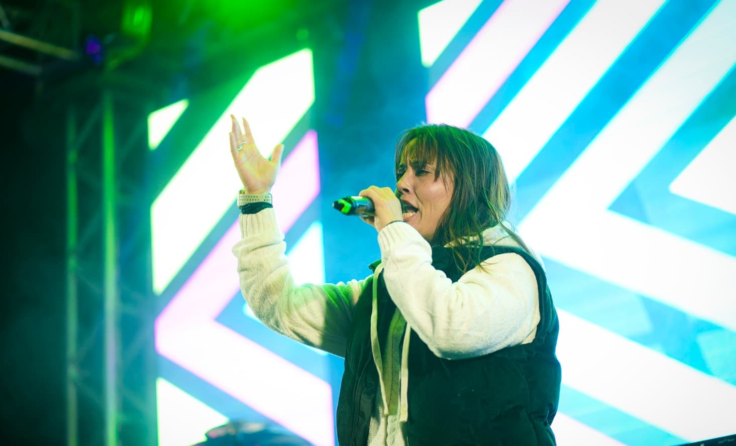 Cantora de colete preto cantando ao microfone com a mão erguida diante do painel de LED