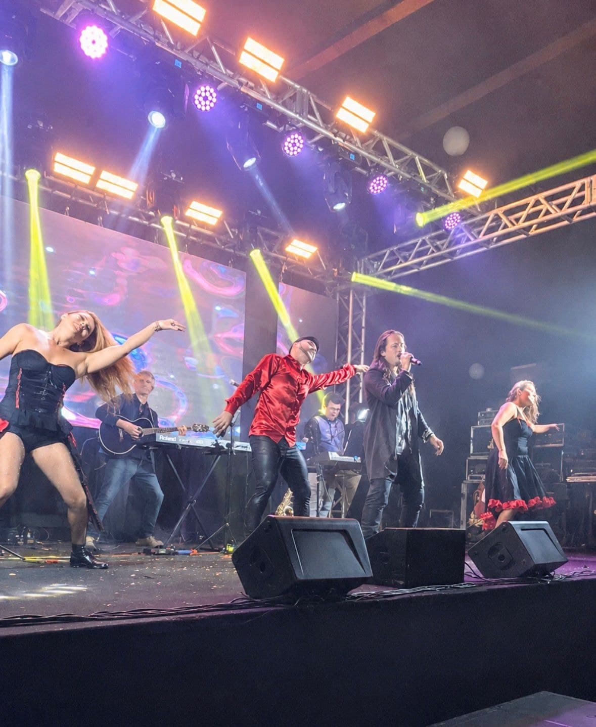 Banda completa no palco com onze integrantes sob iluminação robotizada e telão de LED