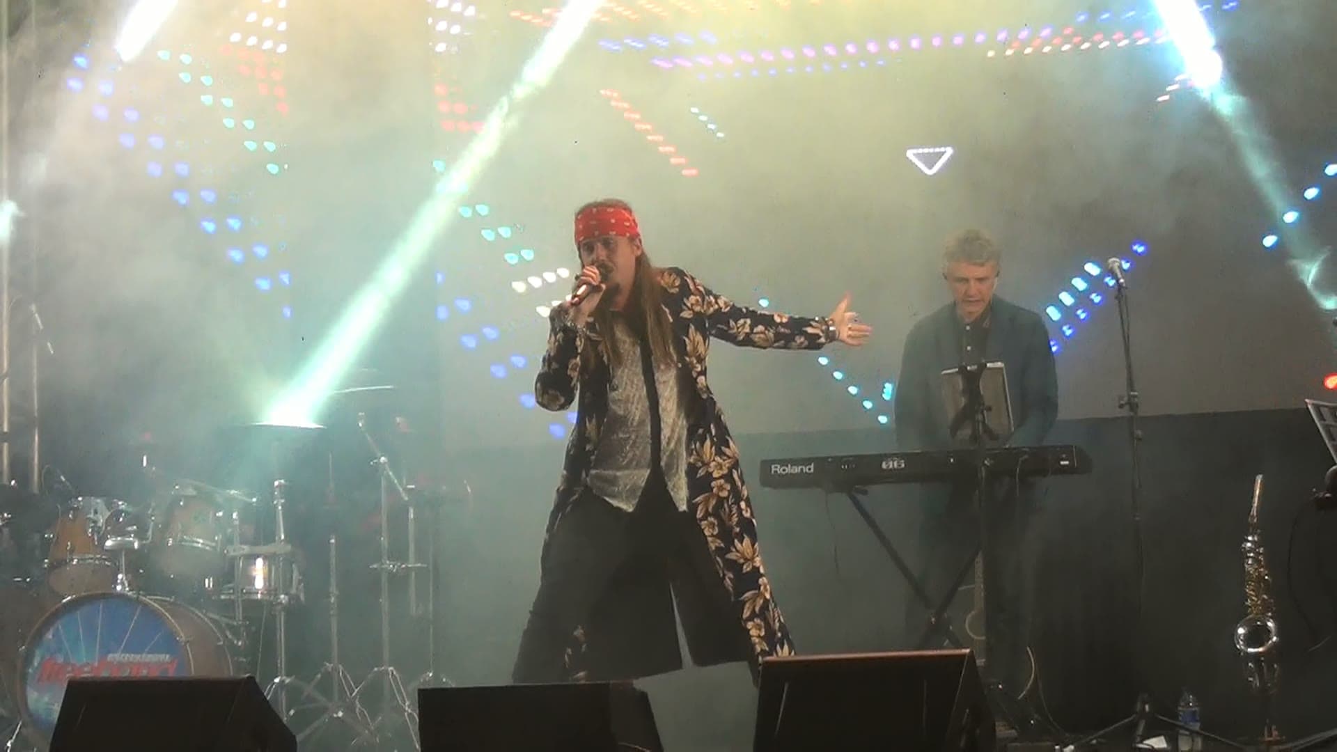 Vocalista de bandana vermelha cantando de braço aberto ao lado do tecladista, entre fachos de luz, fumaça e os pontos do painel de LED