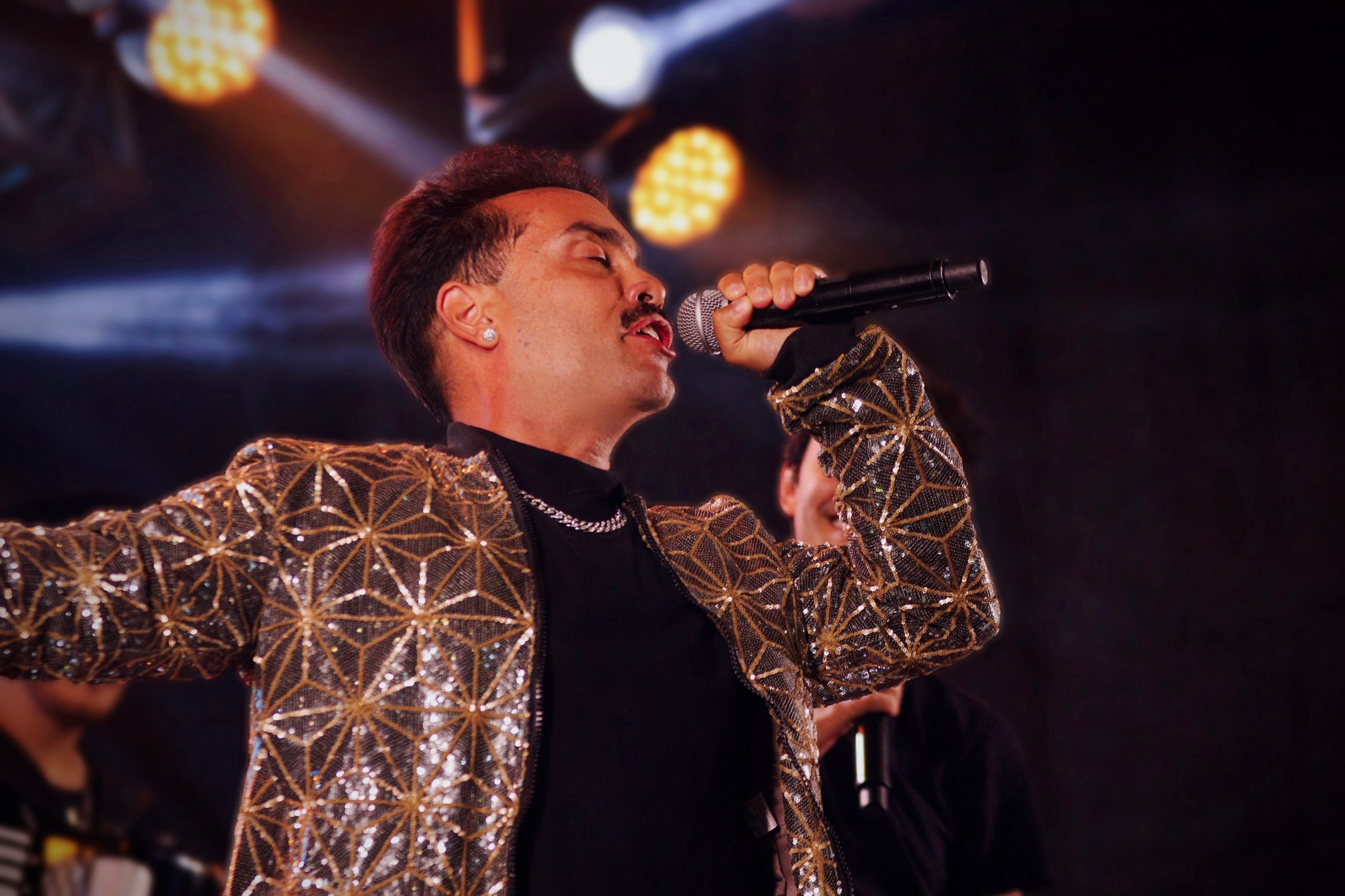 Dois vocalistas da Internacional Freeband cantando ao microfone, um de jaqueta dourada bordada, sob a luz de palco